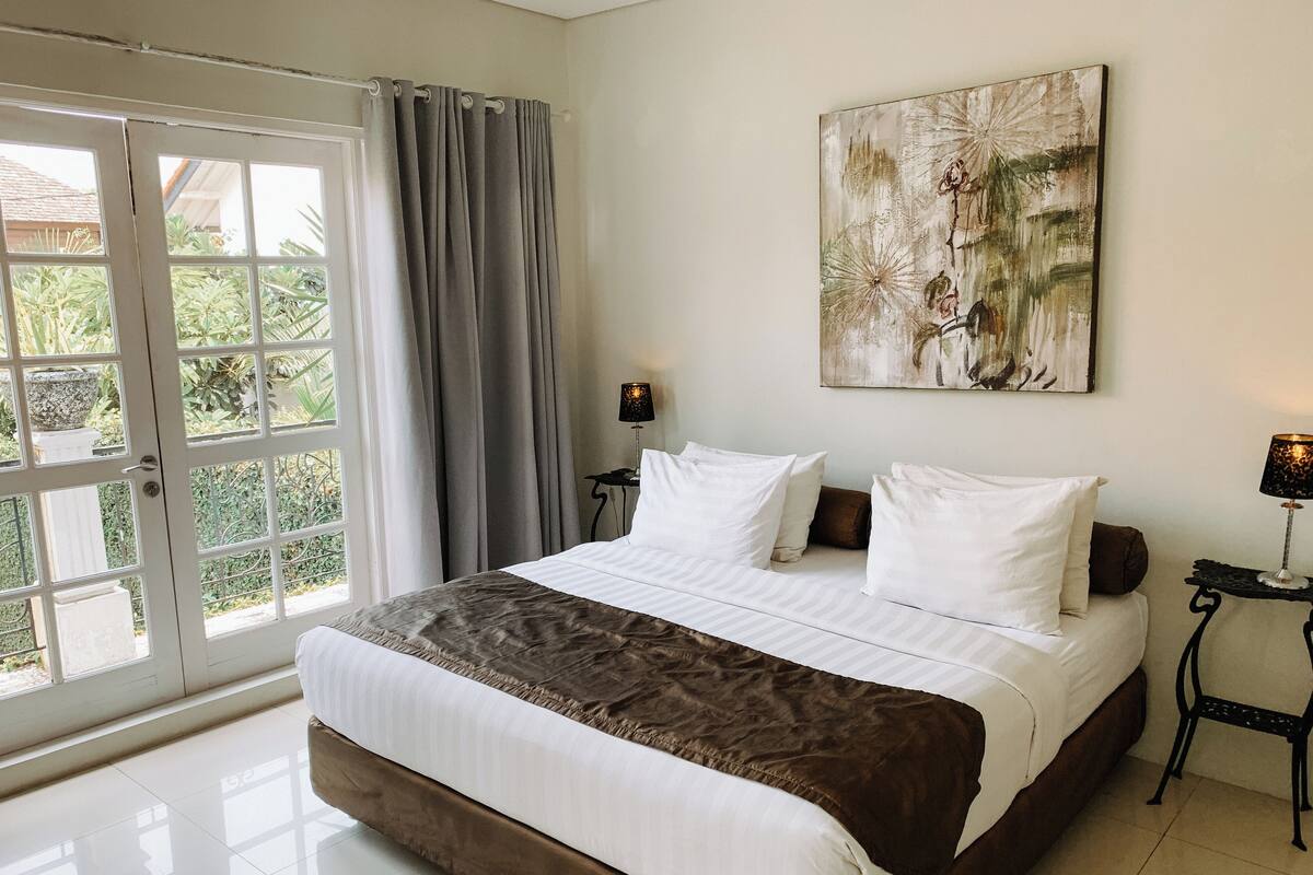 A bedroom with a large bed, a view of greenery through a sliding door, a painting on the wall, and a nightstand with a lamp.