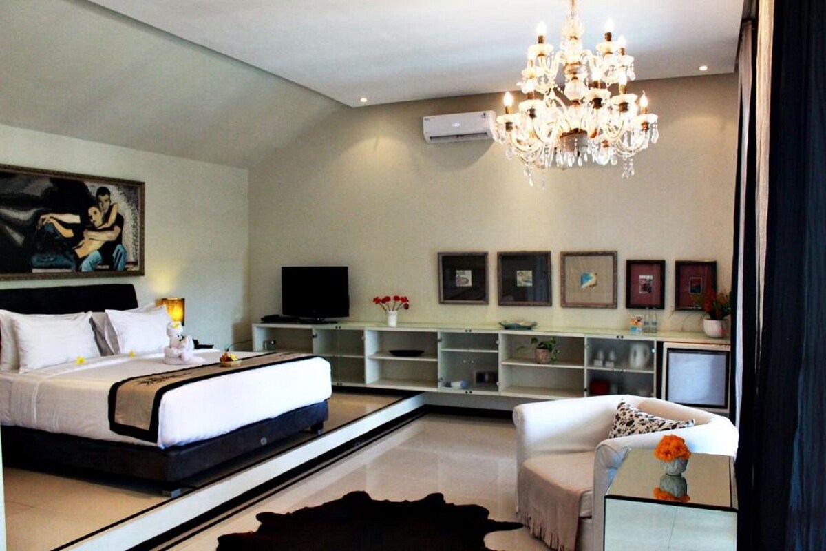 A modern hotel room with a bed, a television, a chandelier, and a desk with a computer.