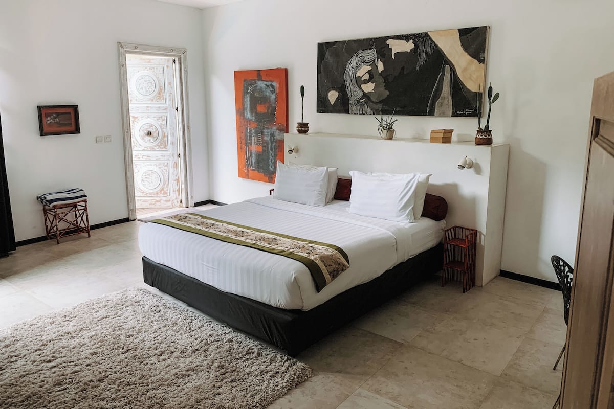 A bedroom with a bed, a door, a small stool, a rug, and a painting on the wall.
