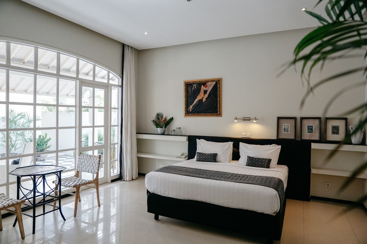 A bedroom with a large bed, a small table, two chairs, a window with curtains, and a wall-mounted picture.