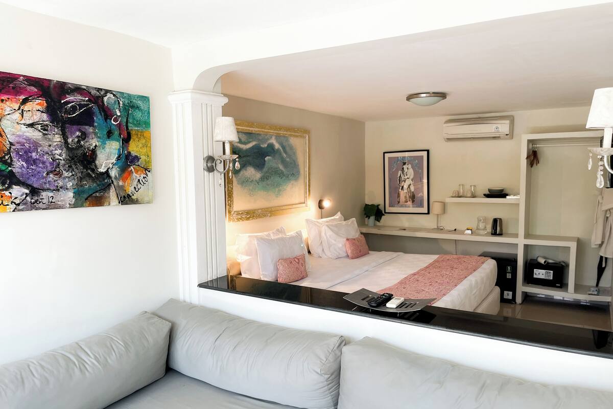 A modern hotel room with a bed, a sofa, a painting, a mirror, and a small kitchenette.