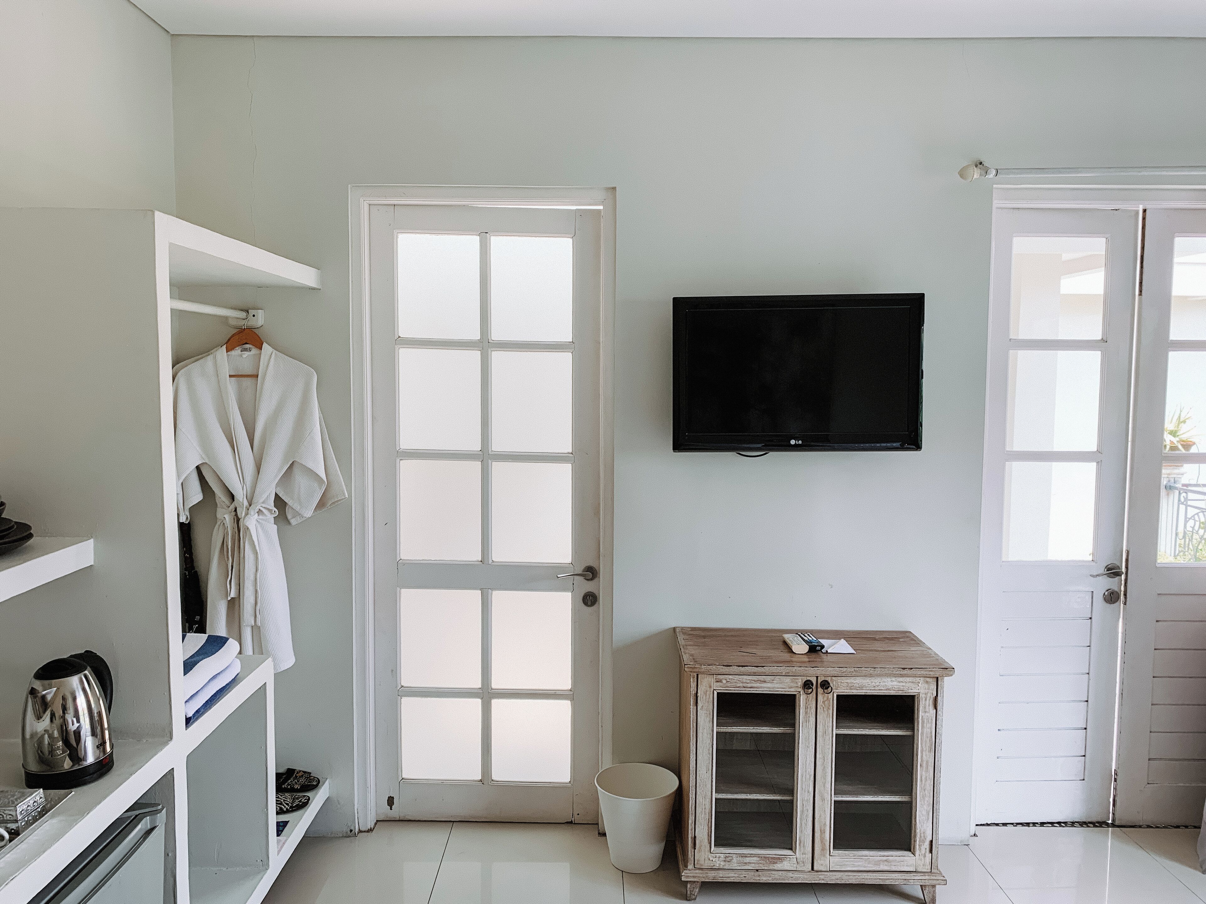Une pièce avec une porte blanche, un téléviseur à écran plat fixé au mur, un meuble en bois et une robe blanche suspendue à un porte-manteau.