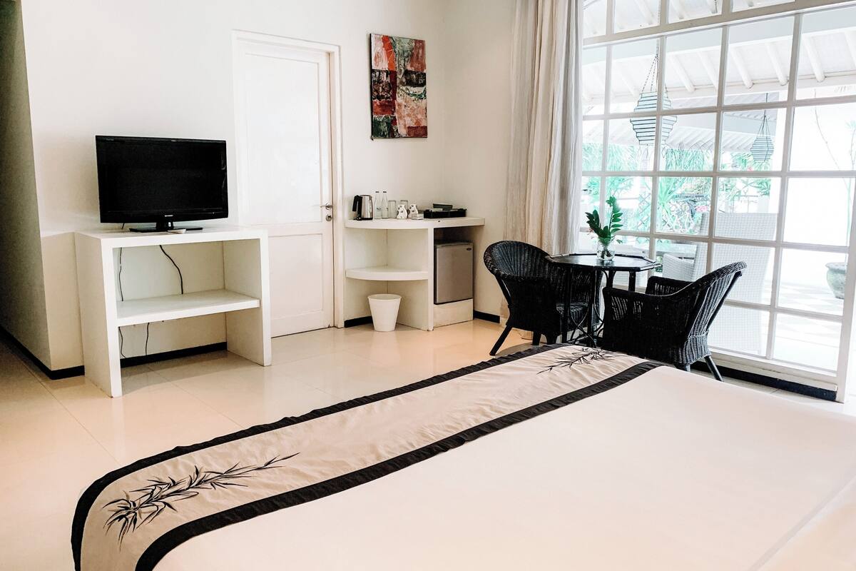 A hotel room with a bed, a TV on a white stand, a small table with chairs, and a window with a view of greenery.