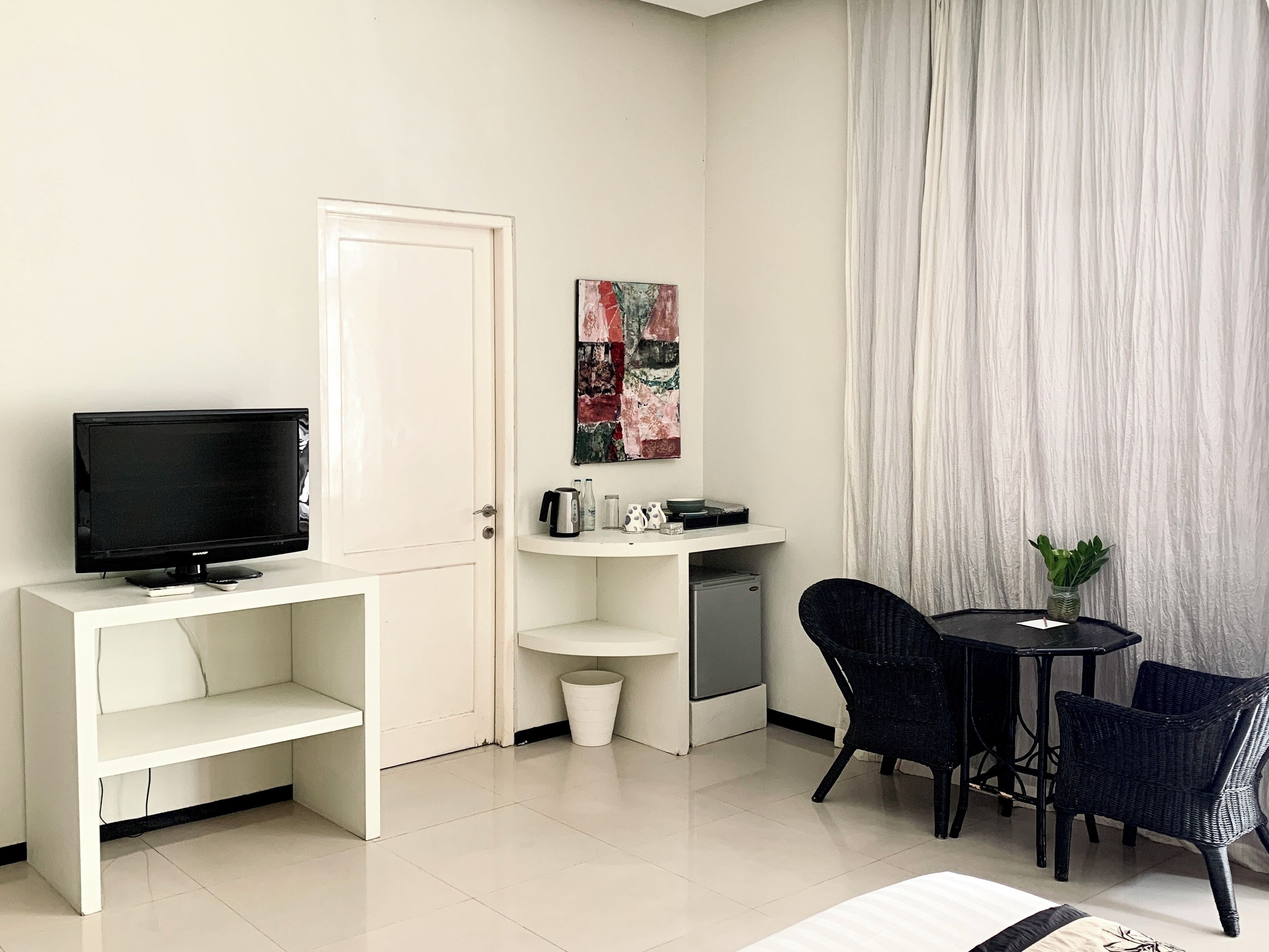 Een compacte kamer met een televisie op een witte standaard, een kleine koelkast, een wit plankje, een schilderij, een tafel met stoelen en een gordijn.