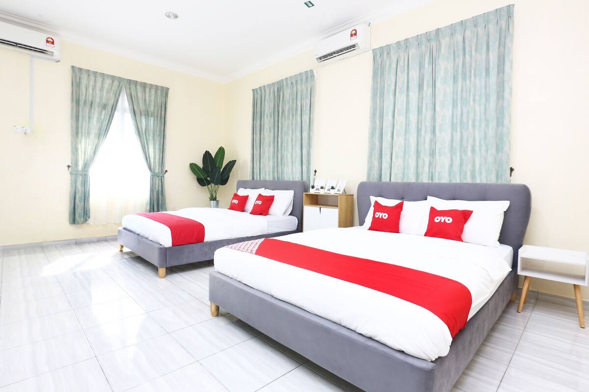 Habitación de hotel con dos camas, cada una con cabecero gris y ropa de cama blanca adornada con almohadas rojas. Se observan unidades de aire acondicionado en las paredes, una planta pequeña sobre una mesita de noche y una mesita auxiliar blanca.