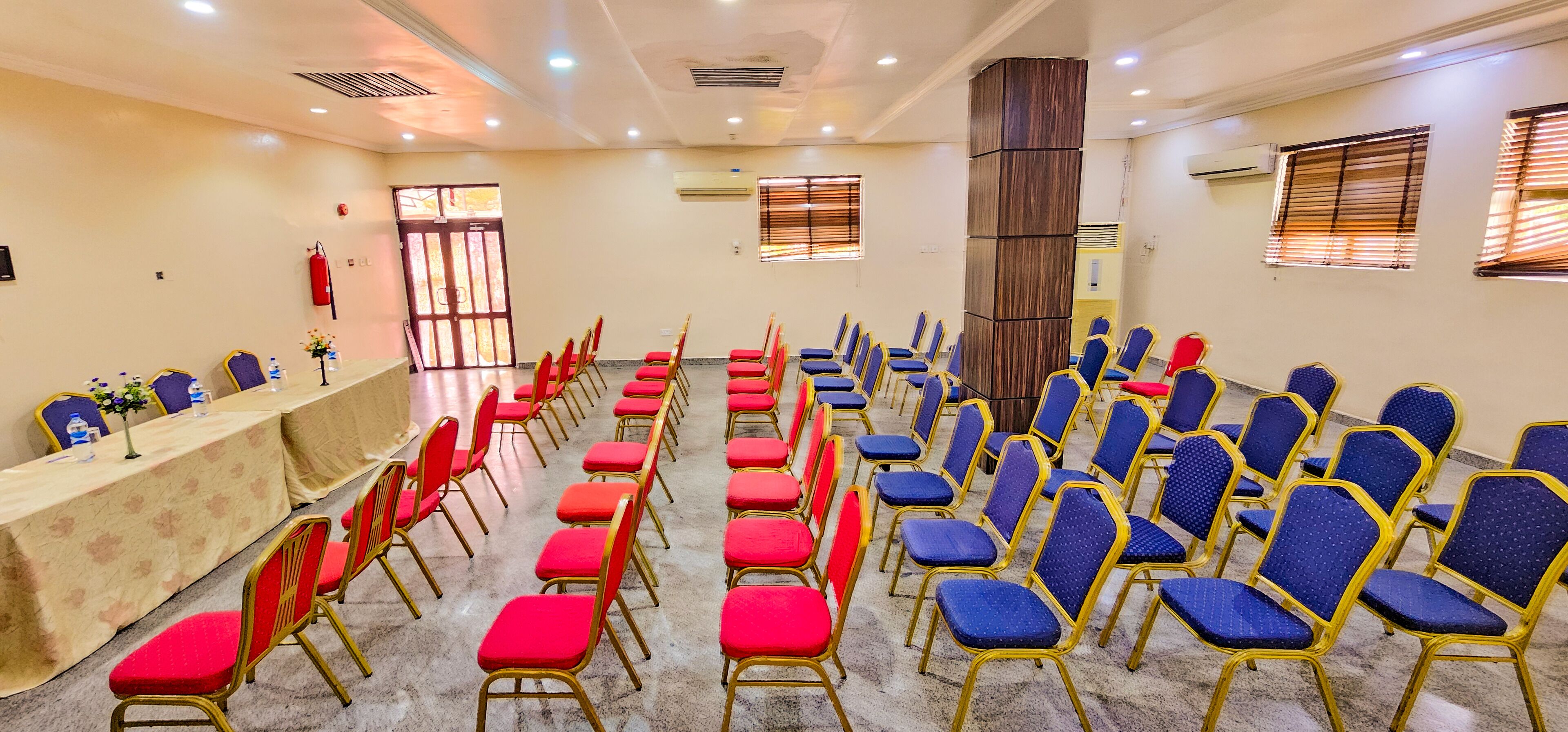 Une salle de conférence avec des rangées de chaises, une longue table agrémentée de décorations florales et des unités de climatisation fixées aux murs.