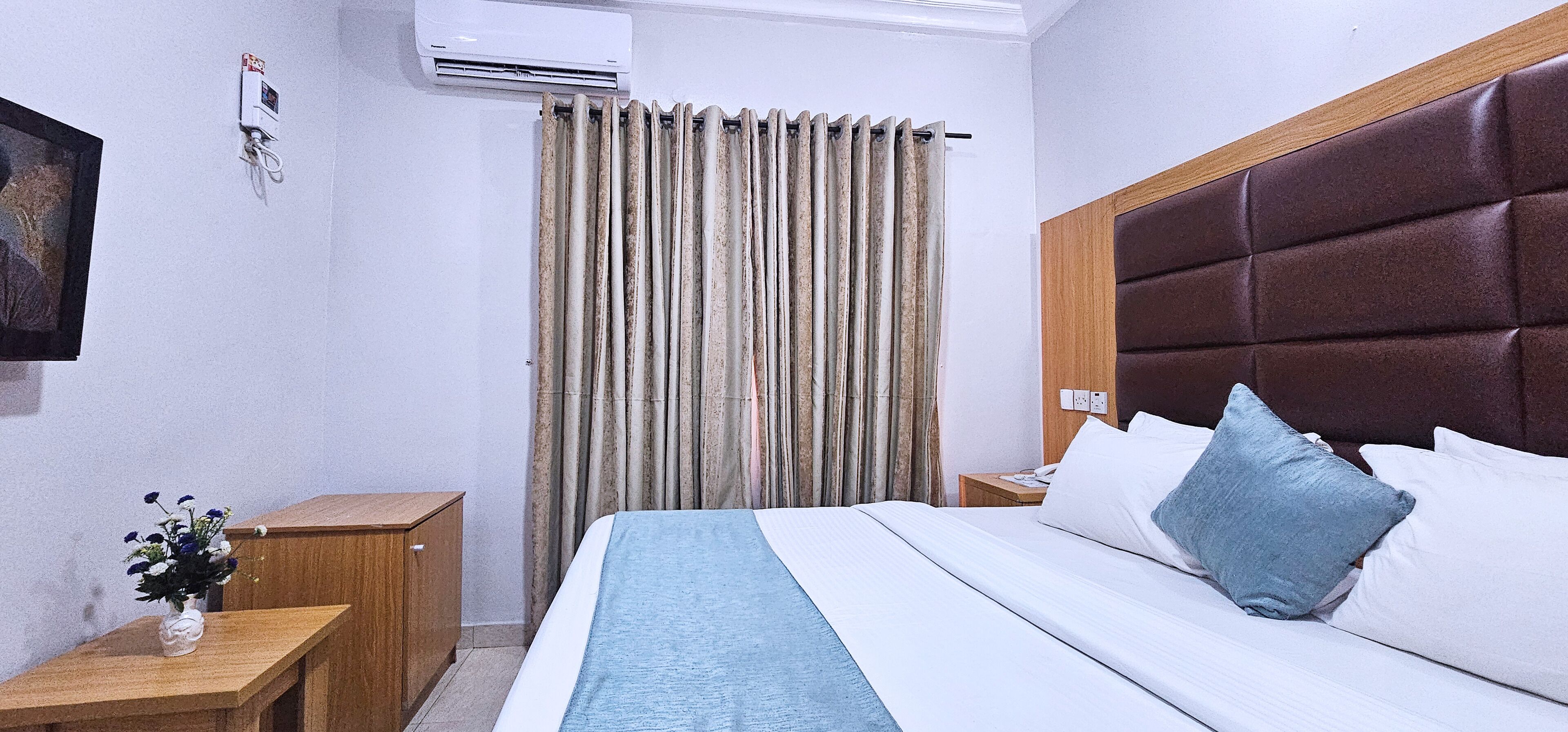 Habitación de hotel con cama, mesita de noche, armario de madera, aire acondicionado de pared y cortina.