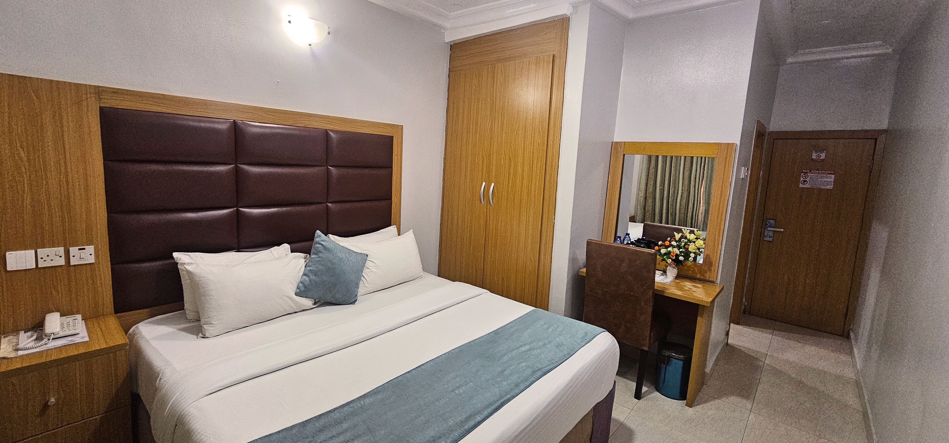 A hotel room with a large bed, a wooden headboard, a desk with a chair, a mirror, and a door.