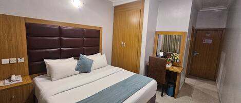 A hotel room with a large bed, a wooden headboard, a desk with a chair, a mirror, and a door.