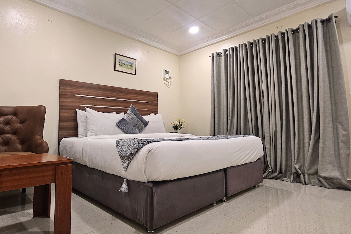 A hotel room with a bed, a wooden chair, a bedside table, a wall-mounted air conditioner, and a window with curtains.