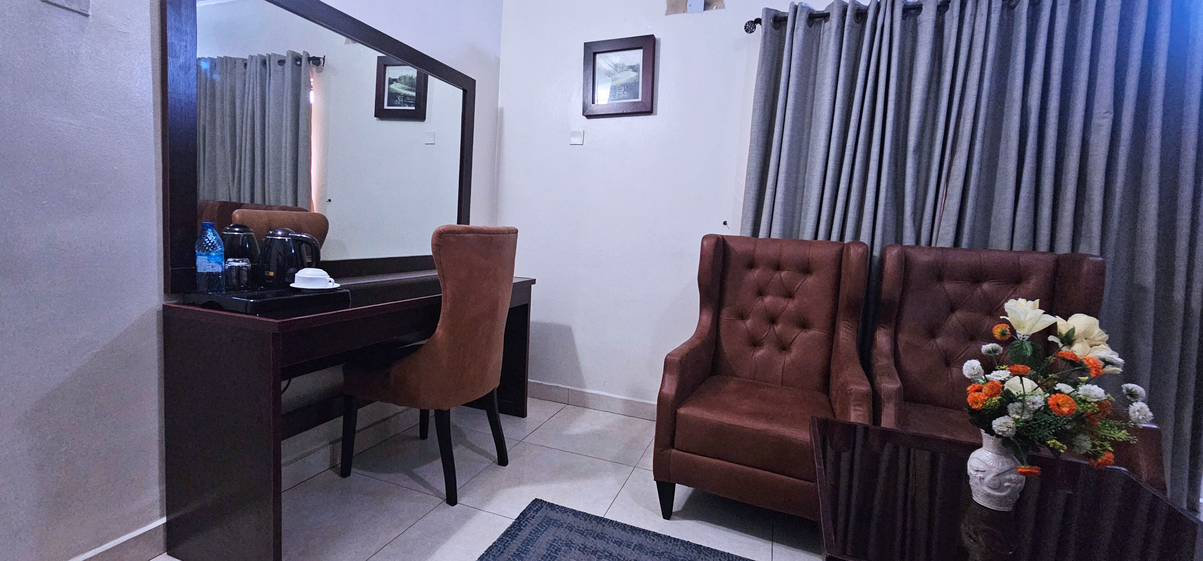 A room with a dark wooden desk, a brown chair, a tufted brown armchair, a mirror, and a vase with flowers.