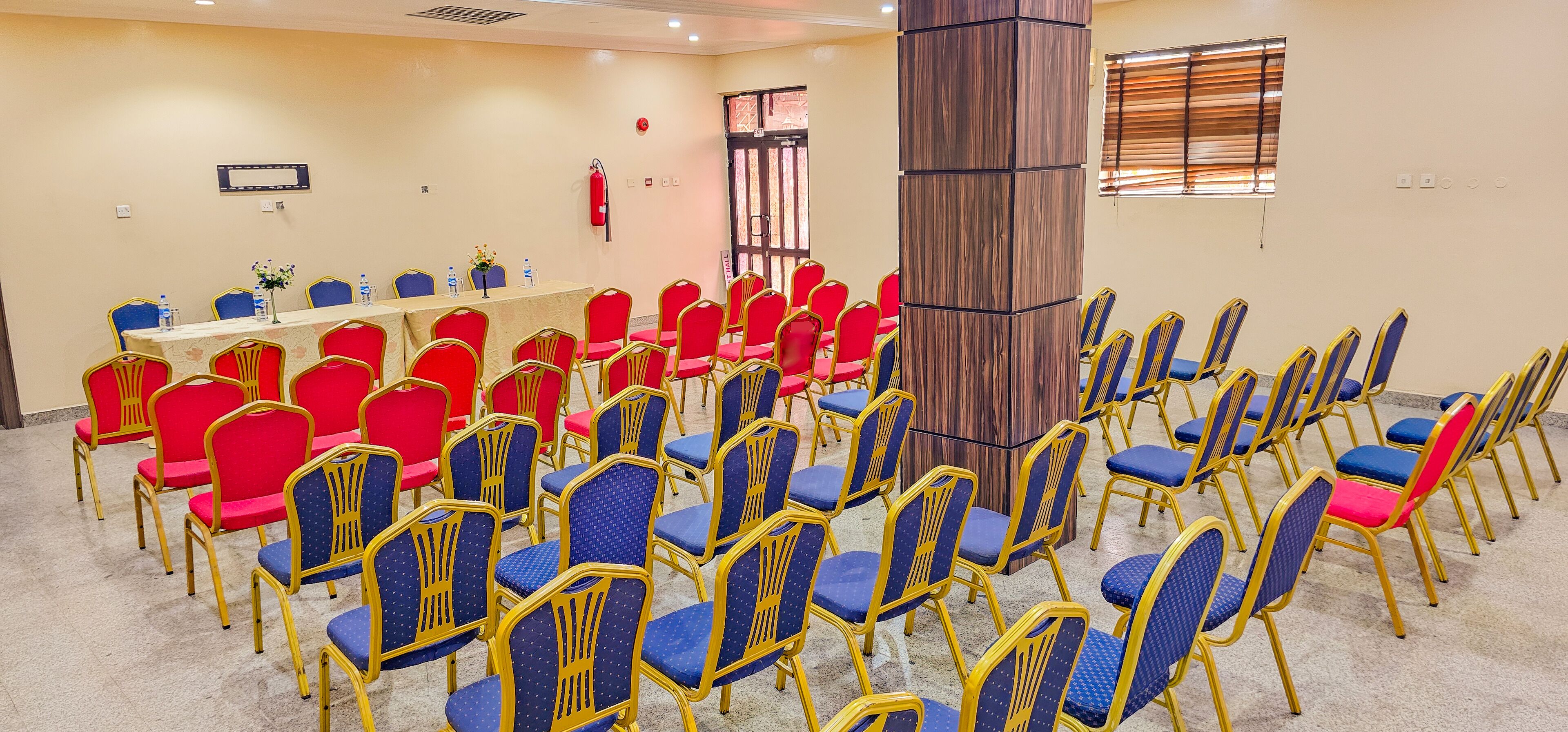 A conference room with rows of red and blue chairs, a long table with water bottles, and a wooden pillar.