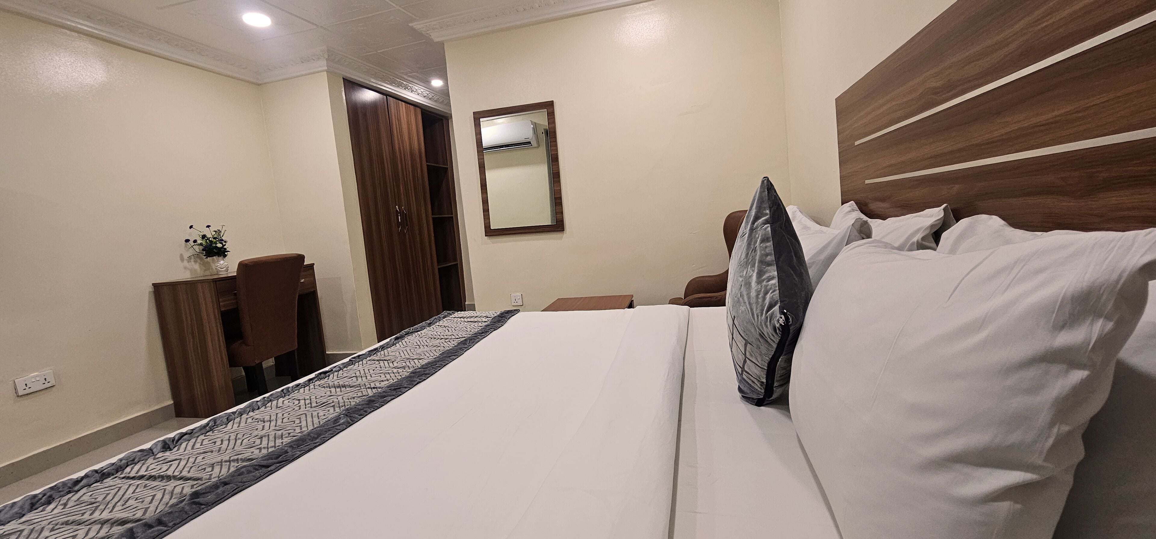 A hotel room with a large bed, a wooden headboard, a wardrobe, a mirror, and a small table with a vase.