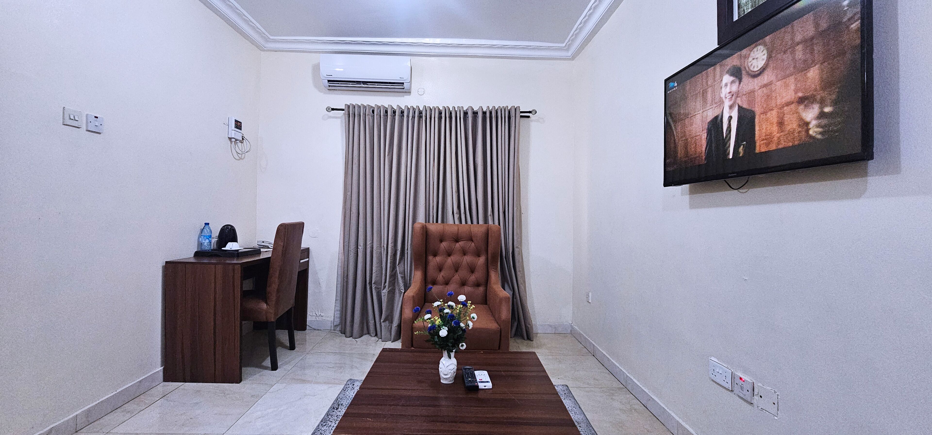 Una sala de estar con un sillón marrón, una mesita de madera, un televisor montado en la pared y un ventanal con cortinas.