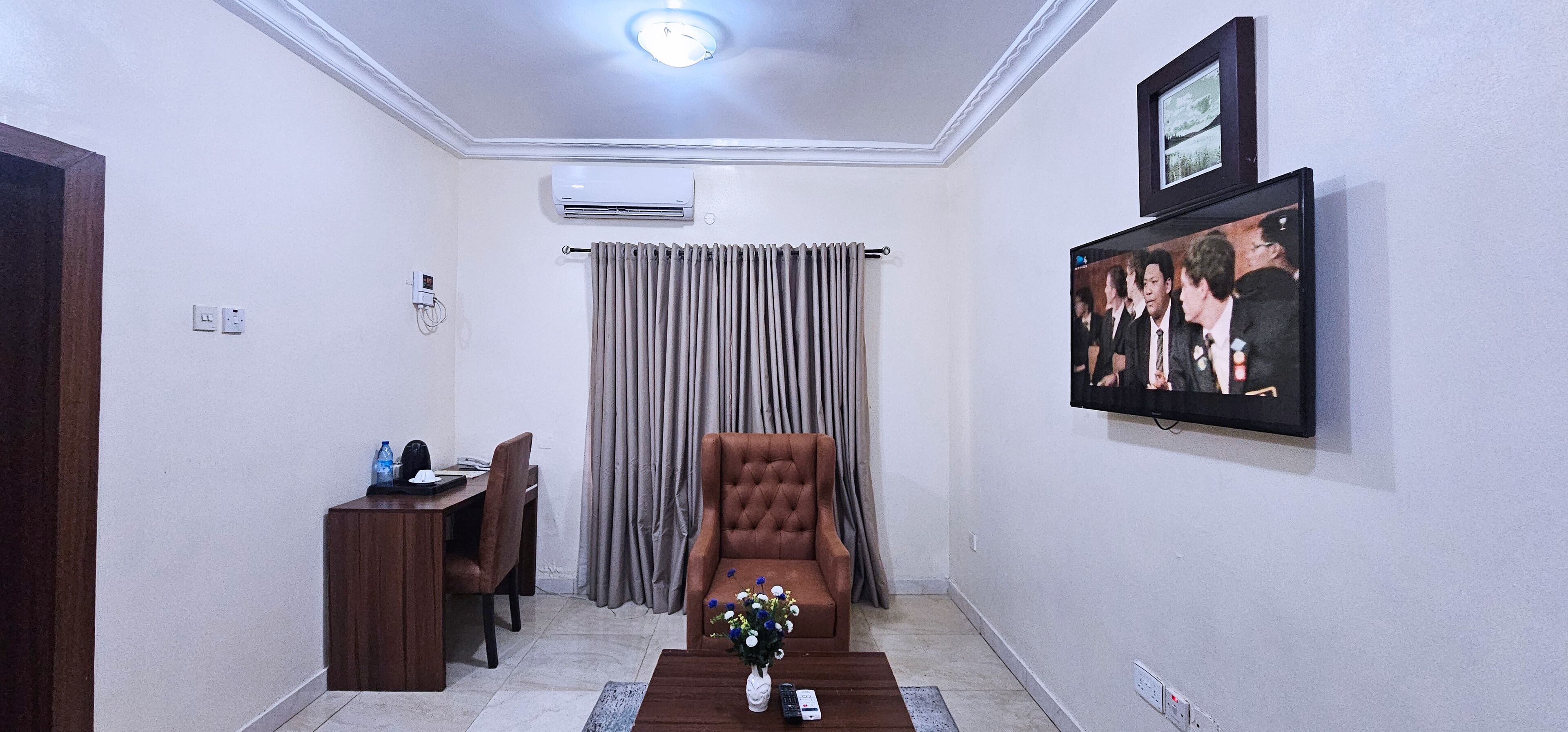 A hotel room with a dining area, a living area with a brown armchair, a TV mounted on the wall, and an air conditioning unit.