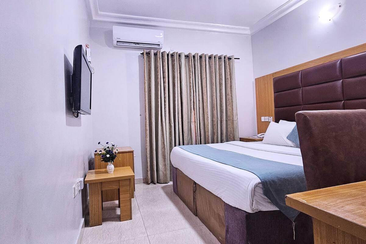 A hotel room with a bed, a desk, a chair, a small table with a vase, a TV mounted on the wall, and a window with curtains.