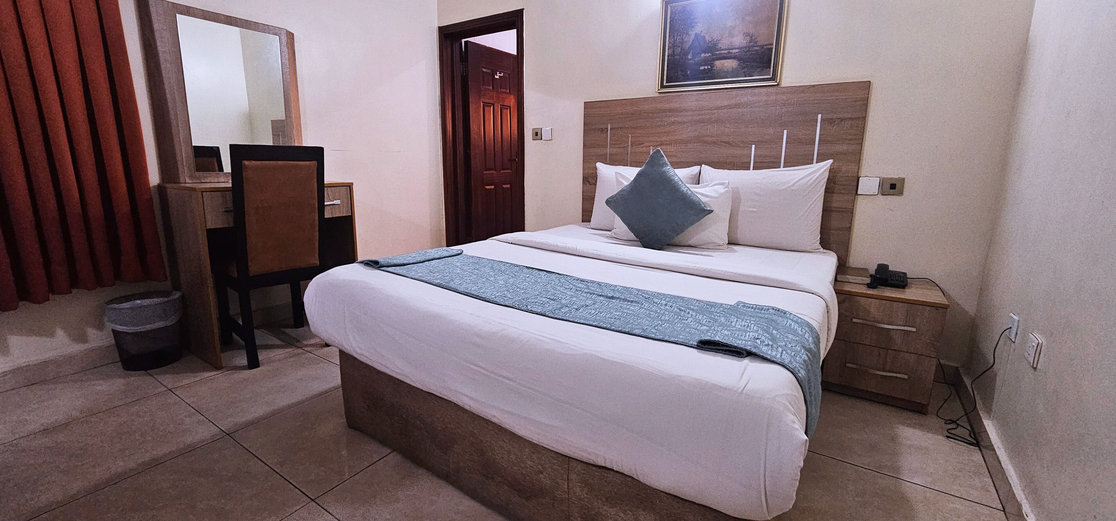 A hotel room with a bed, bedside table, nightstand, wooden headboard, and a mirror.