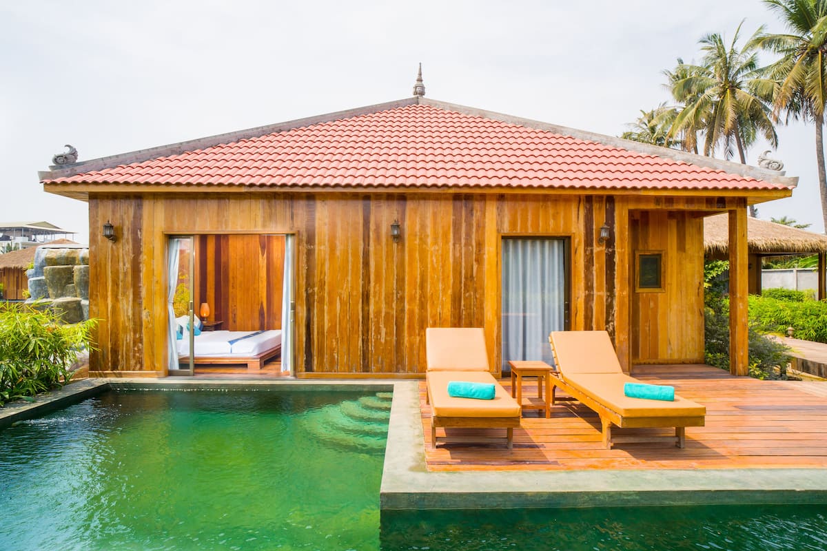 A wooden villa with a red tiled roof, a pool, and two lounge chairs with towels.