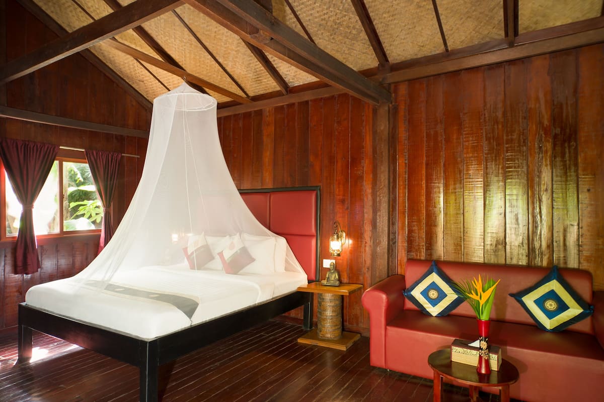 A room with a bed under a mosquito net, a red sofa with decorative pillows, a small table with a vase, and wooden walls.