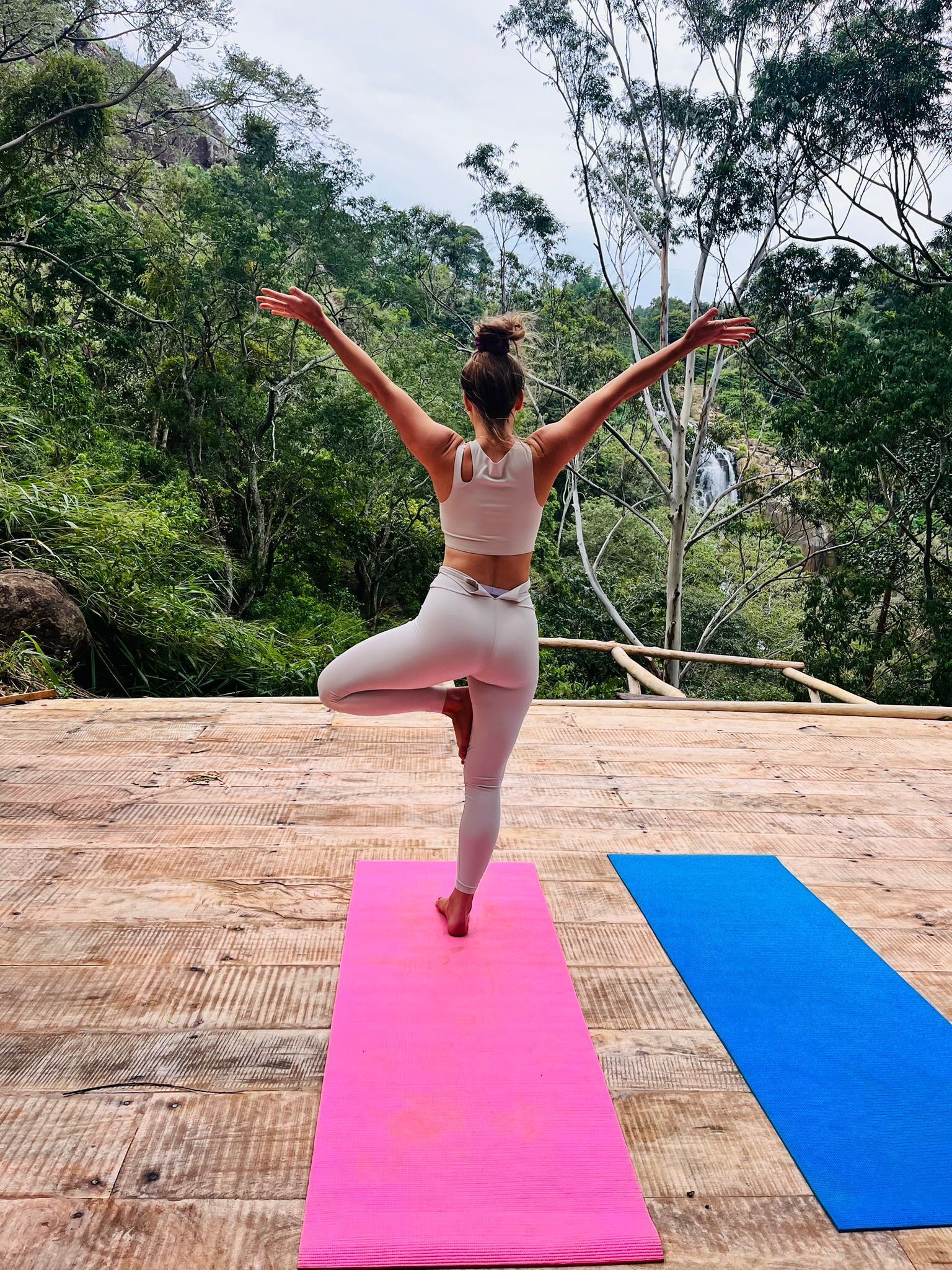 Una persona realiza una postura de yoga sobre una esterilla rosa al aire libre, con un bosque de fondo.