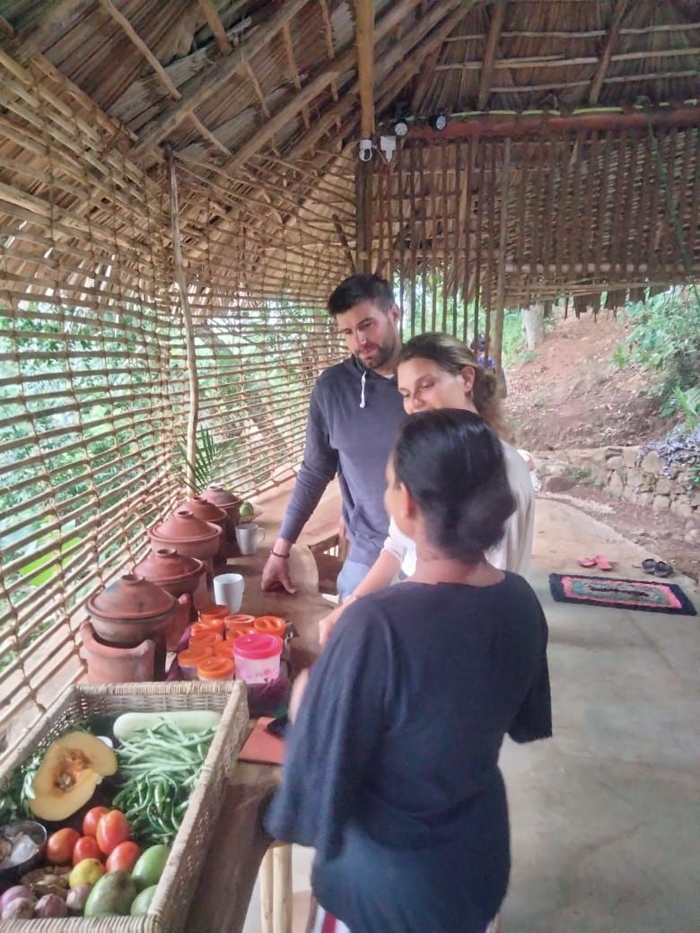 Una estructura rústica de bambú con techo de paja, una mesa con macetas de barro y productos frescos, y un sendero de piedra al fondo.