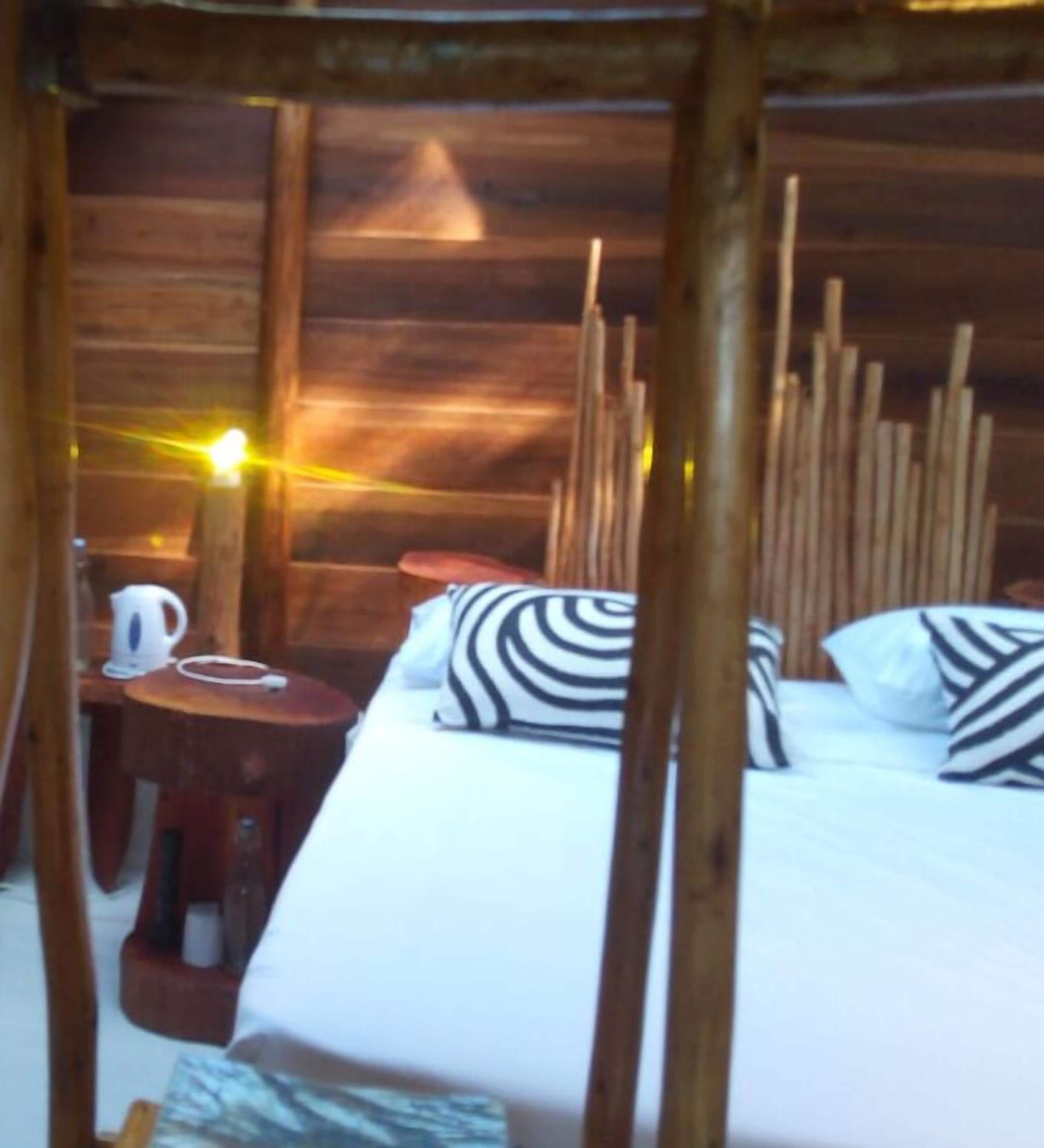 Habitación de cabaña de madera con una cama, almohadas a rayas, una mesa con un hervidor y una silla.