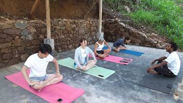 Un grupo de personas sentadas sobre esterillas de yoga al aire libre, con un muro de piedra y follaje en el fondo.