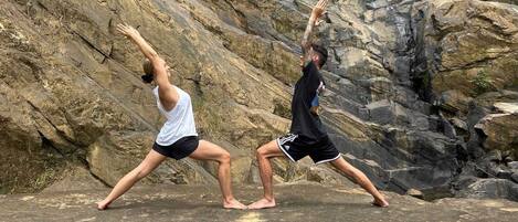 Two individuals performing a yoga pose on a rocky surface with a mountainous backdrop.