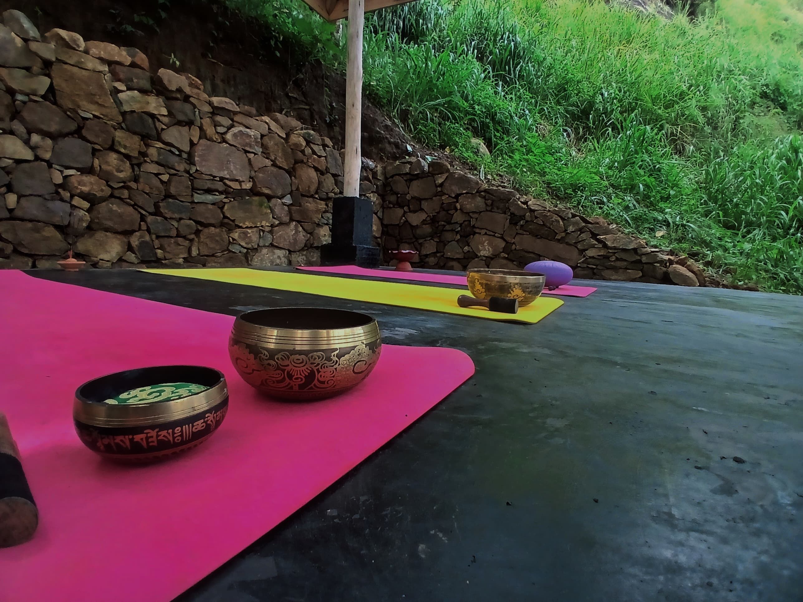 Un espace dédié à la méditation, avec deux bols de méditation posés sur des tapis roses, un mur de pierre et, en arrière-plan, une colline verdoyante.
