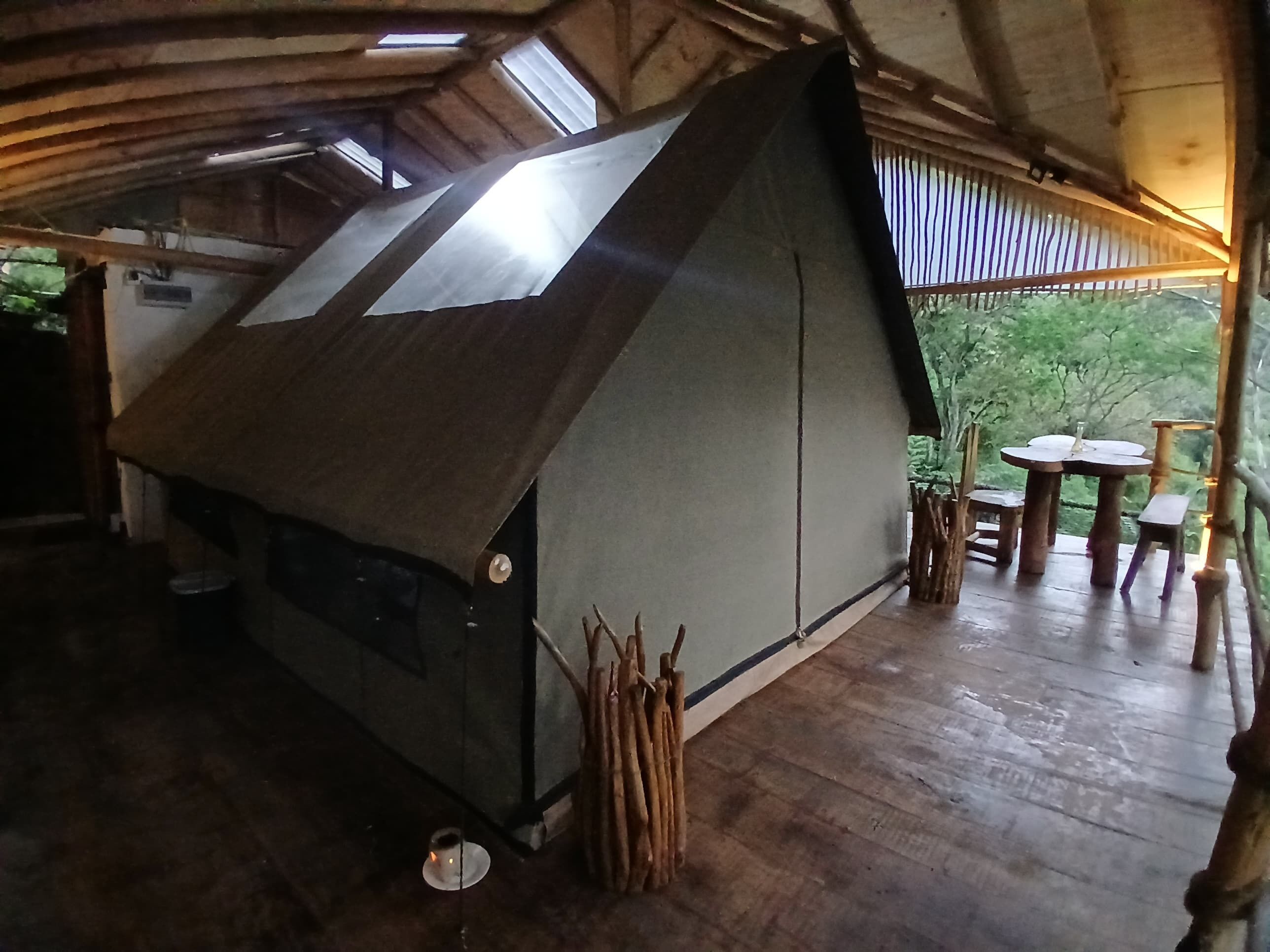 Una estructura rústica tipo tienda con techo inclinado, suelo de madera y una pequeña mesa con sillas en el exterior.