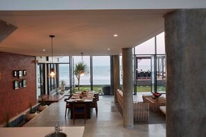 A spacious dining area with a view of the ocean, featuring a long table set for dining, a brick wall, and large windows. - Brand new absolute beachfront retreat (east selemadeg)