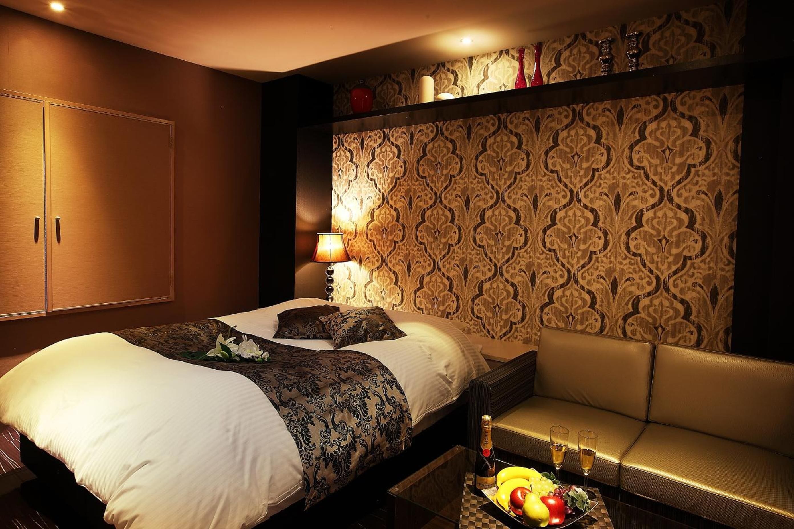 A hotel room with a large bed, a sofa, a table with fruit and drinks, and a patterned wallpaper.
