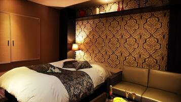 A hotel room with a large bed, a sofa, a table with fruit and drinks, and a patterned wallpaper.