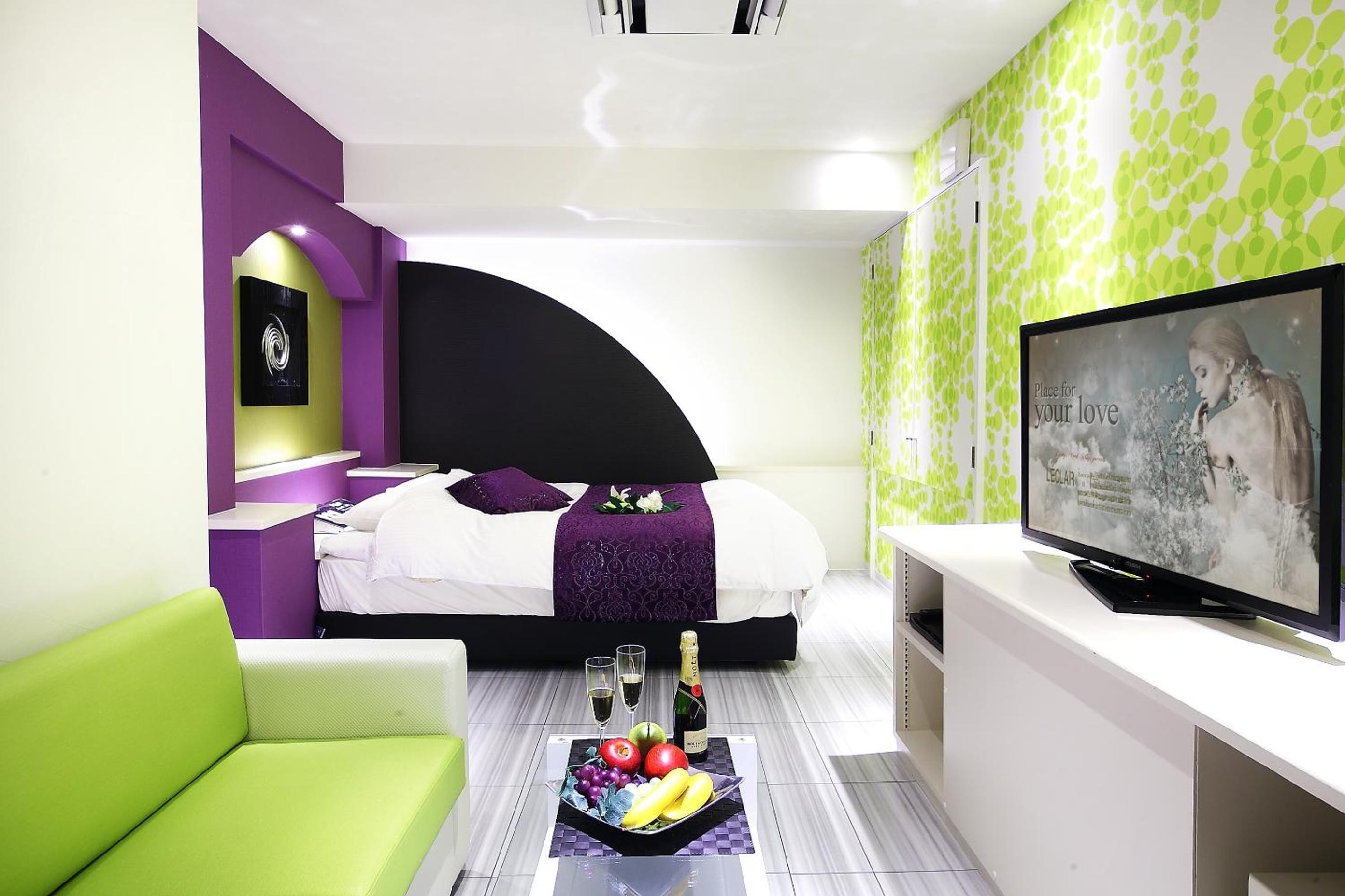 Una habitación de hotel moderna con una cama, un sofá verde, un televisor y una bandeja con frutas y champán en el suelo.