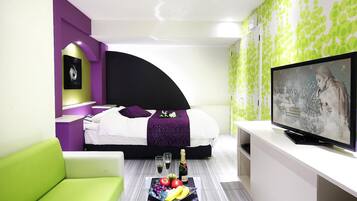 Una habitación de hotel moderna con una cama, un sofá verde, un televisor y una bandeja con frutas y champán en el suelo.