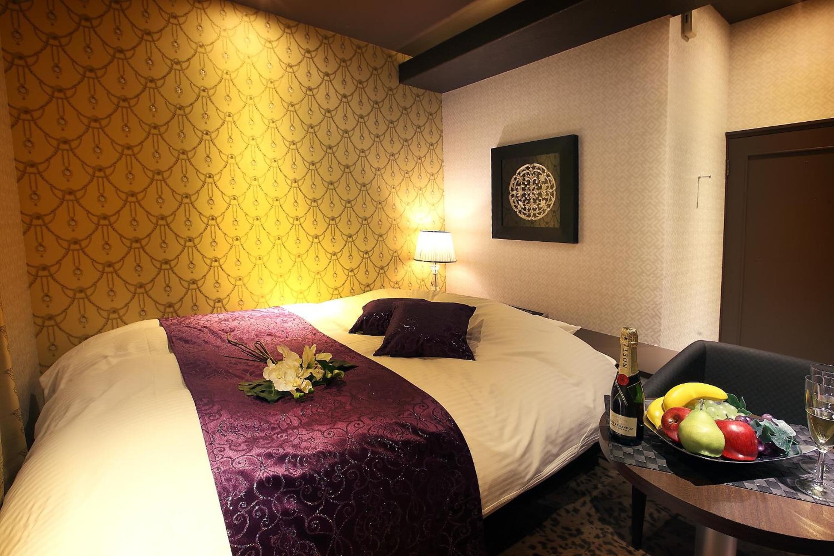 Een hotelkamer met een bed, een nachtkastje met champagne, een schaal fruit en een lijst met kunstwerk aan de muur.