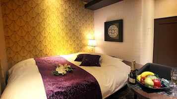 Een hotelkamer met een bed, een nachtkastje met champagne, een schaal fruit en een lijst met kunstwerk aan de muur.