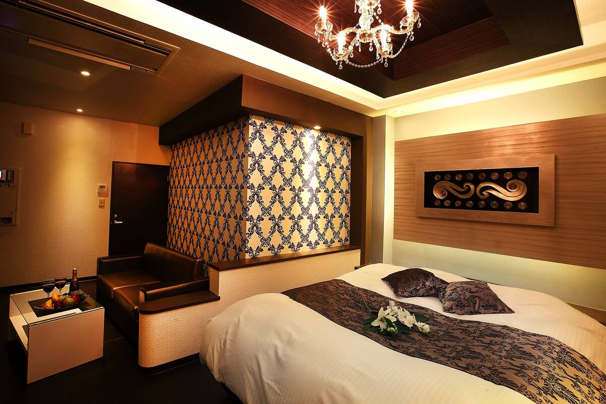 A modern hotel room with a bed, a small seating area, a decorative wall, and a chandelier.