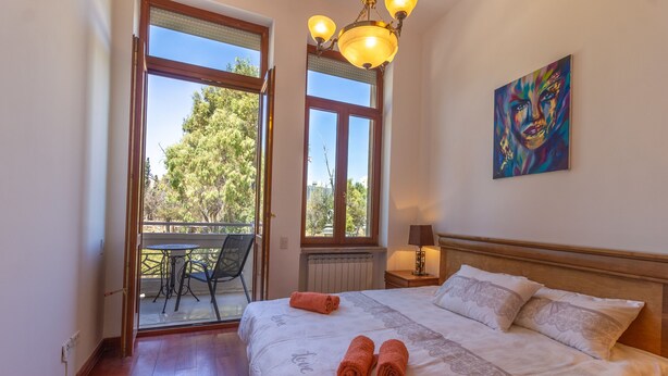 A bedroom with a bed, two pillows, a nightstand, a lamp, a painting, and a view of an outdoor area with a table and chairs. - Art apartment in Mamila (Jerusalem)
