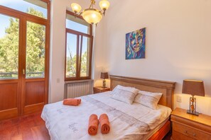 Quarto de hotel com uma cama grande, duas mesas de cabeceira com luminárias, uma janela com vista para árvores e um quadro na parede.
