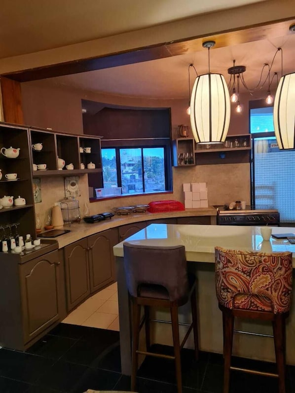 A modern kitchen with a large island, bar stools, and a view of the city through a large window. - Haggai - Tourist Bangalow (Colombo)