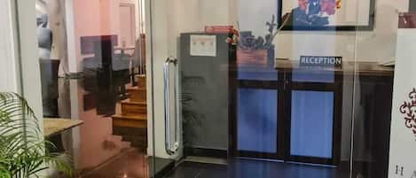 Zona de recepción con puerta de vidrio, mostrador de recepción, escalera y una planta en maceta.