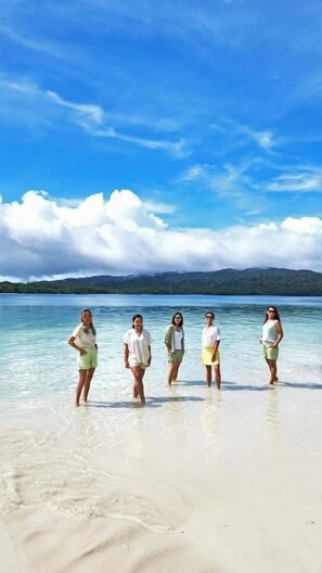 A group of people standing on a beach with clear water and mountains in the background.