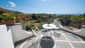 A balcony with a table and chairs, potted plants, and a view of the sea and hills.