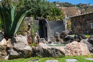 Un jardín de rocas con fuente de agua, que incluye una cascada y un estanque, rodeado de grandes rocas y follaje.