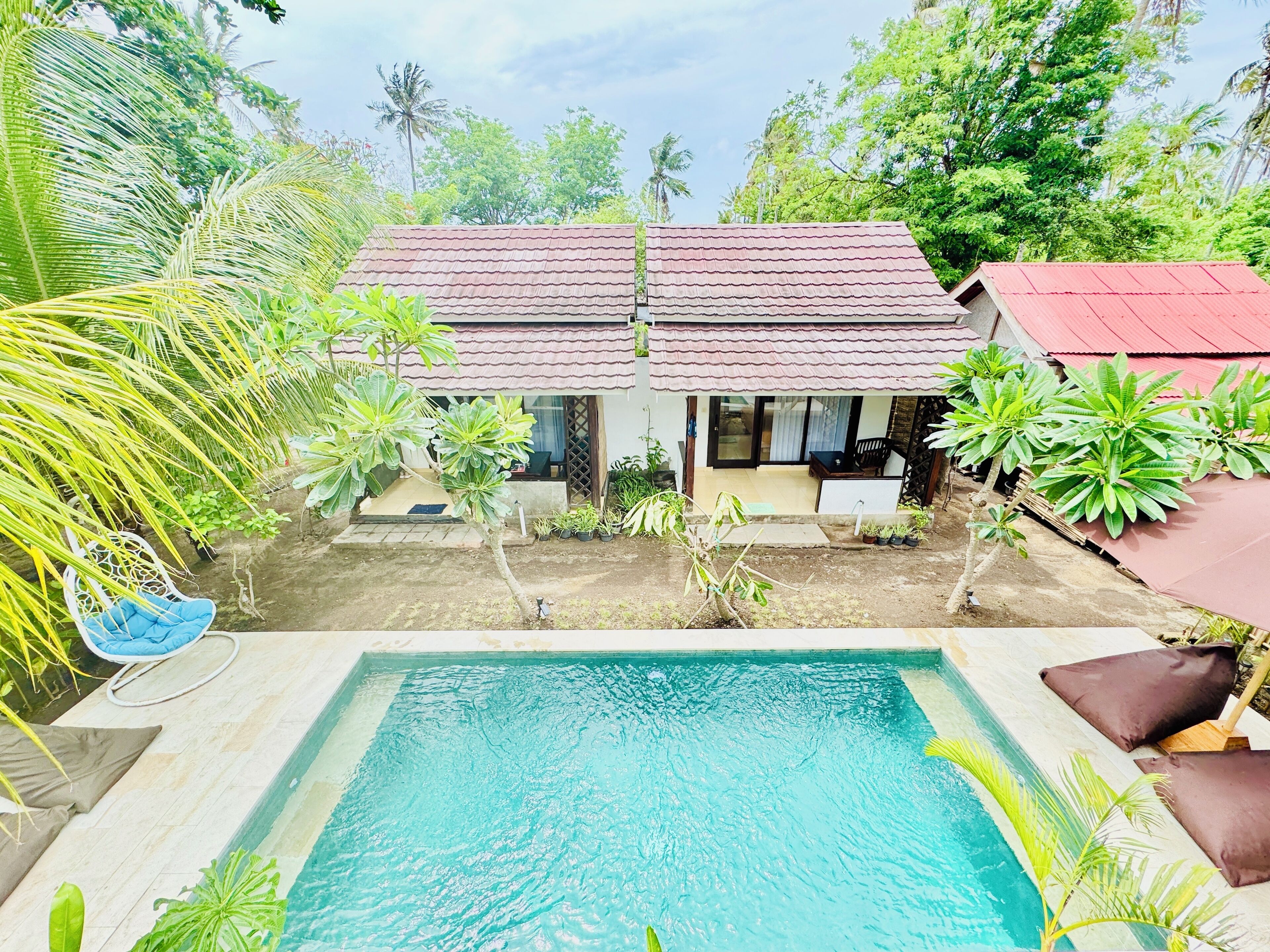 A pool area with two houses, one with a red roof and another with a brown roof, surrounded by greenery and a white fence.