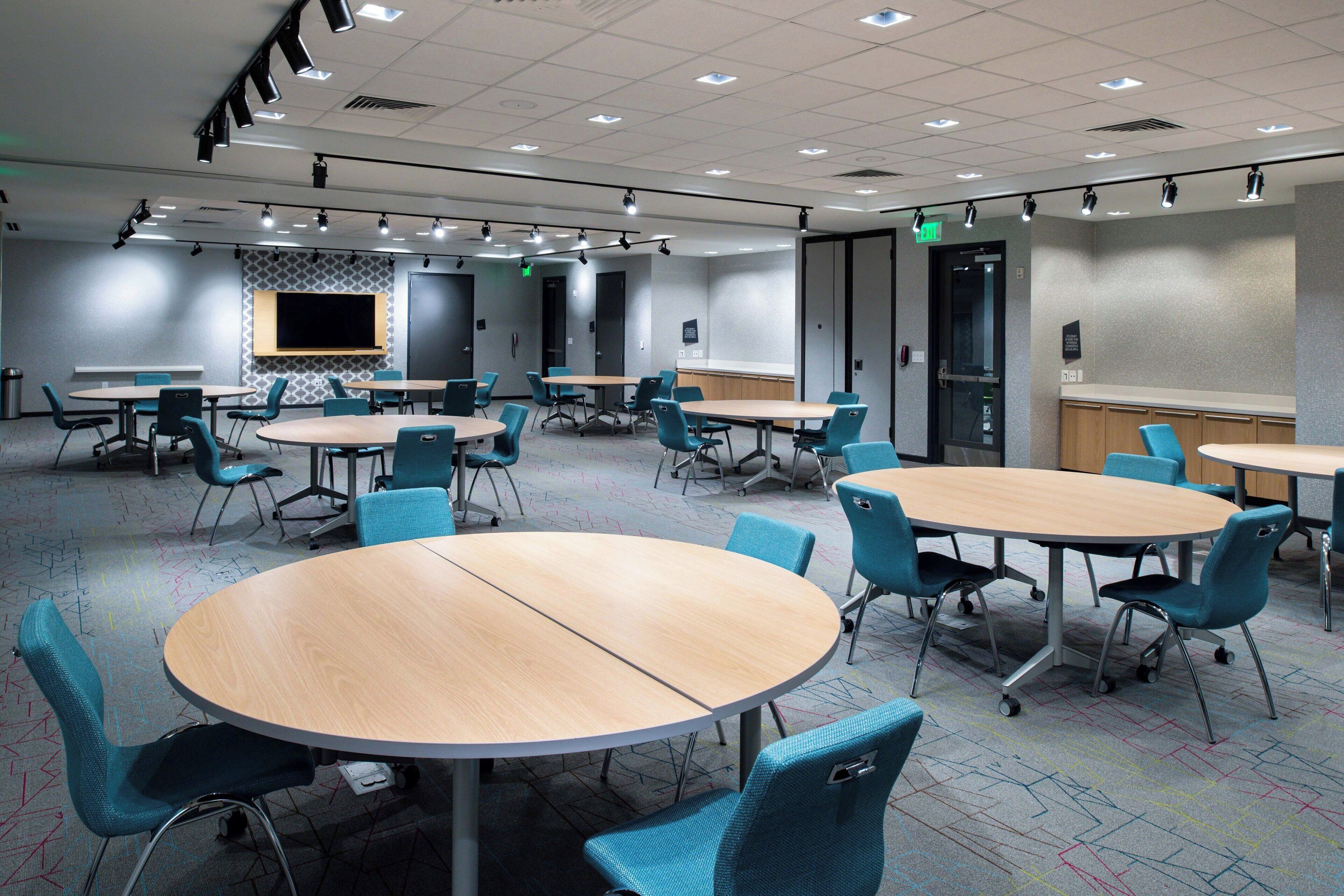A conference room with multiple tables and chairs, equipped with ceiling lights and a large screen.