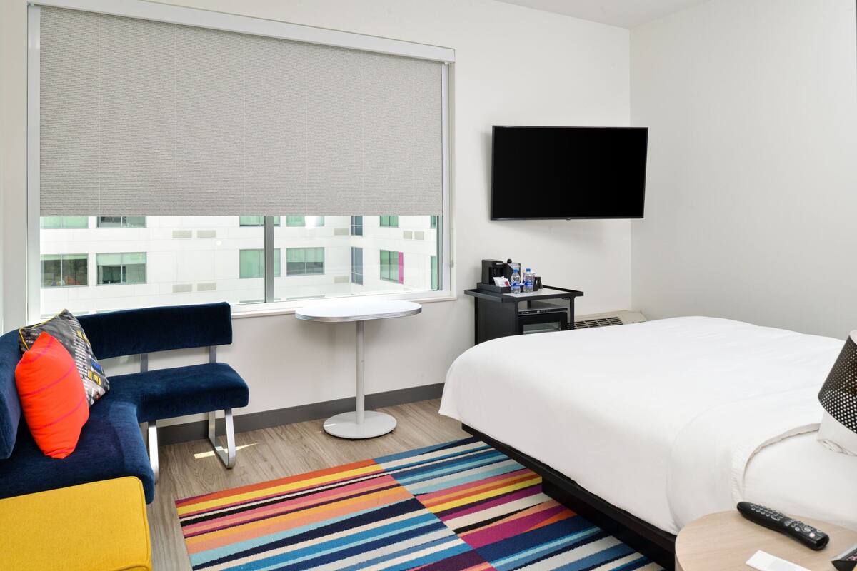 A hotel room with a large window, a bed, a desk, a chair, a small table, and a flat-screen TV.