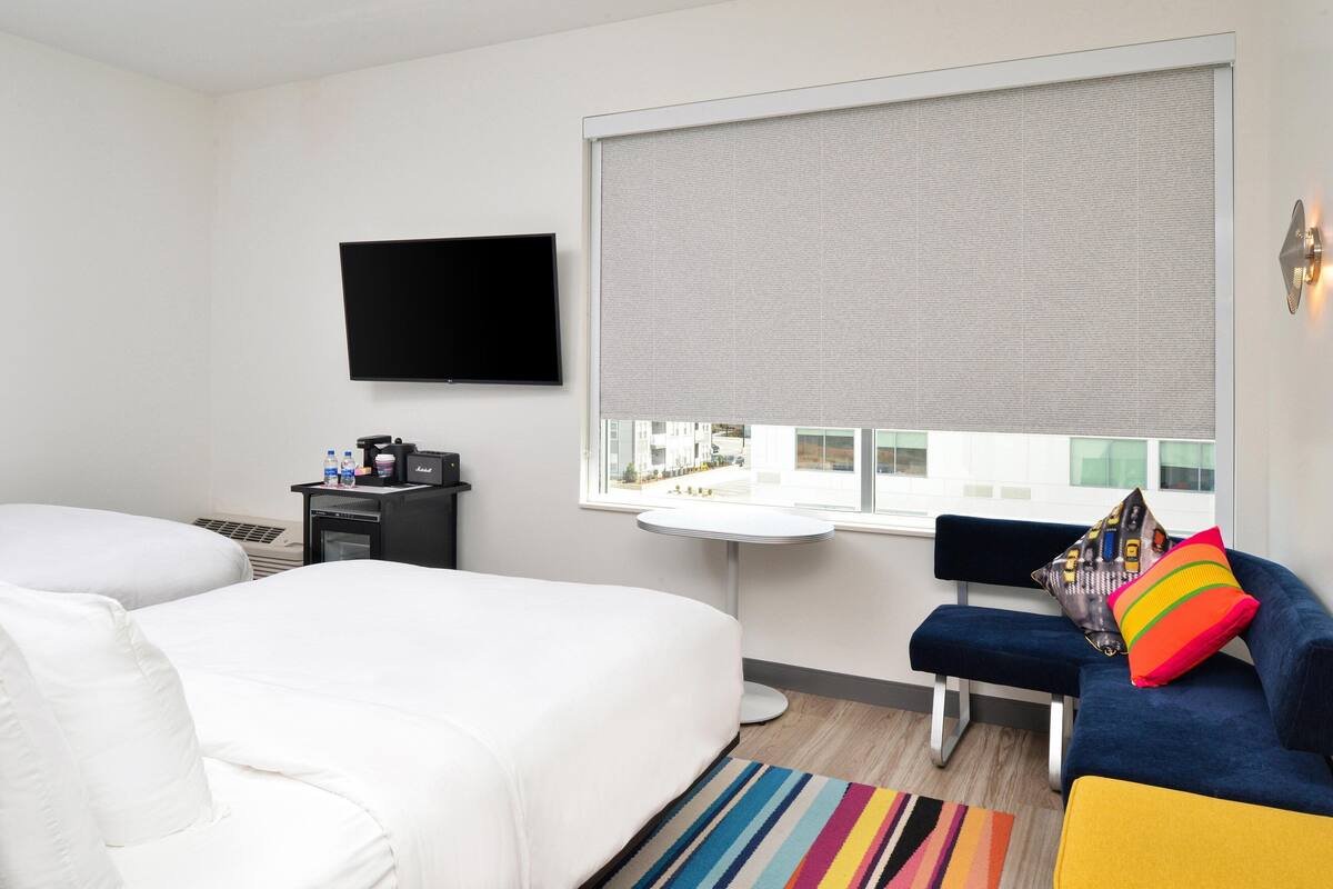 A hotel room with two beds, a TV, a window with blinds, a small table, and a sofa with colorful cushions.