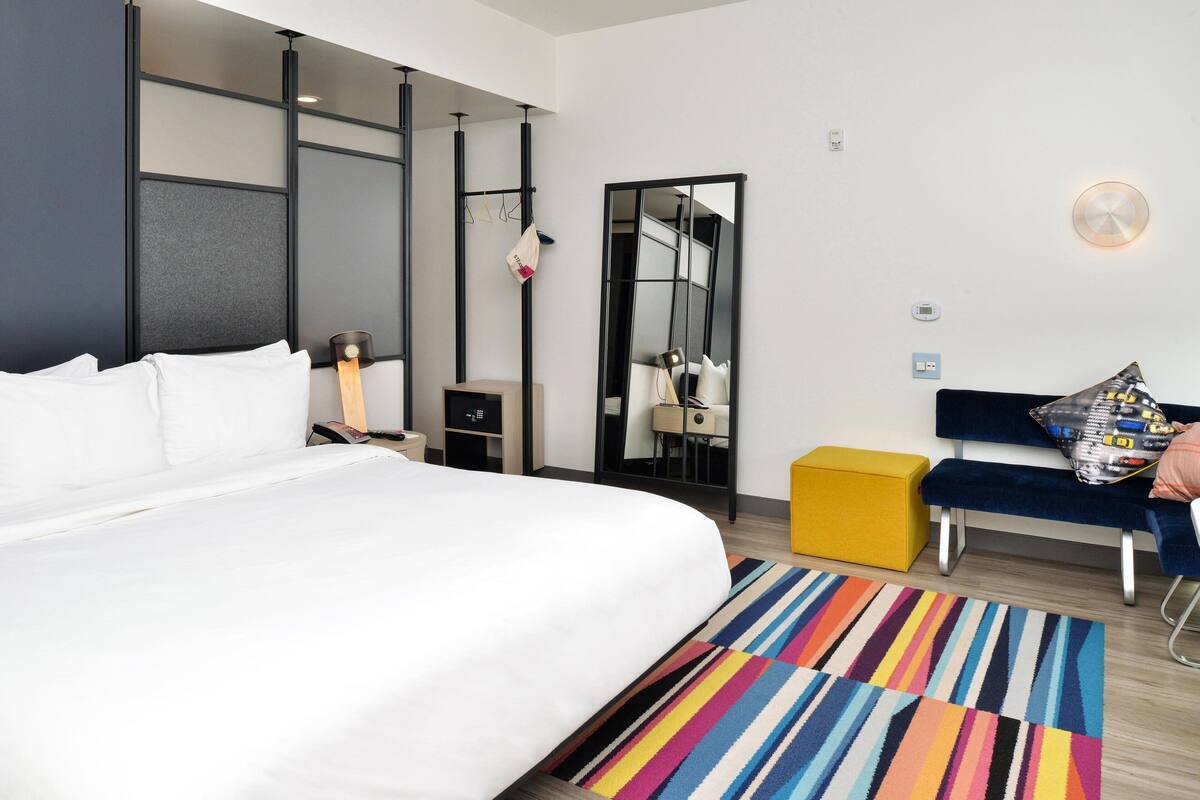 A modern hotel room with a large bed, a small desk, a mirror, a bench, and a colorful striped rug.